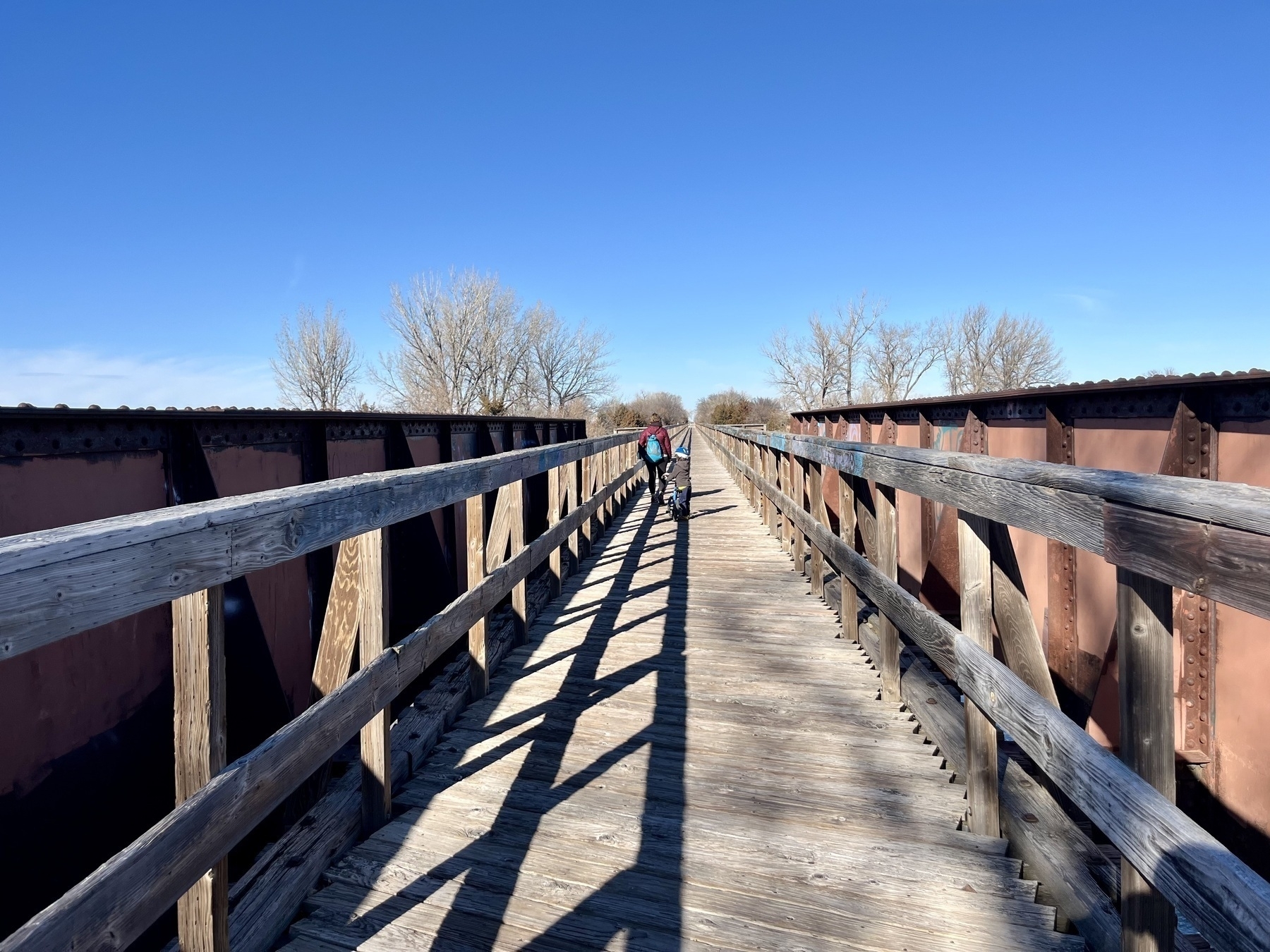 My wife and son walk across a wooden bridge with railings over the Platte river on a sunny day.