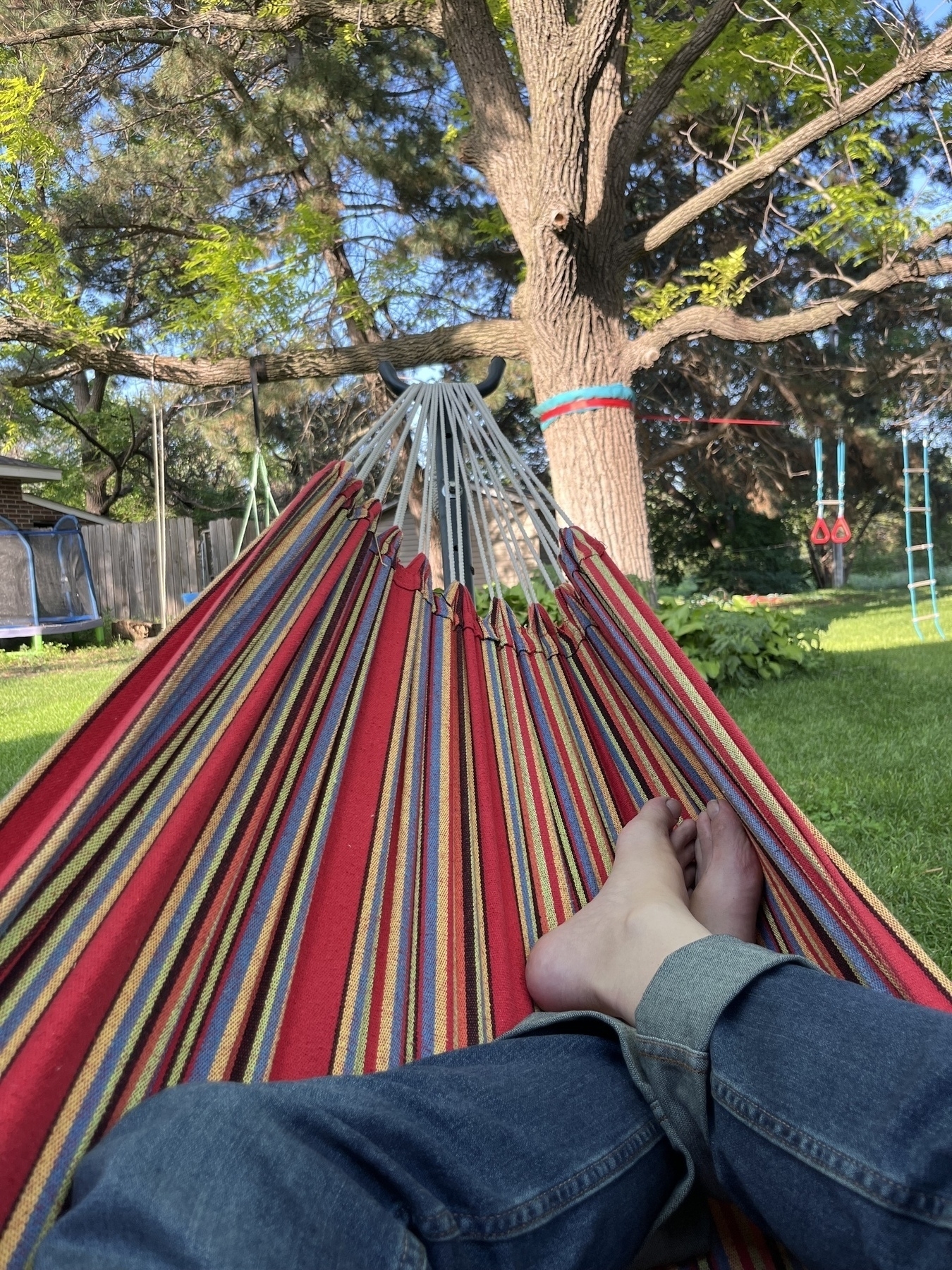 A person relaxes in a colorful hammock in a backyard, surrounded by grass, trees, and a small playground.