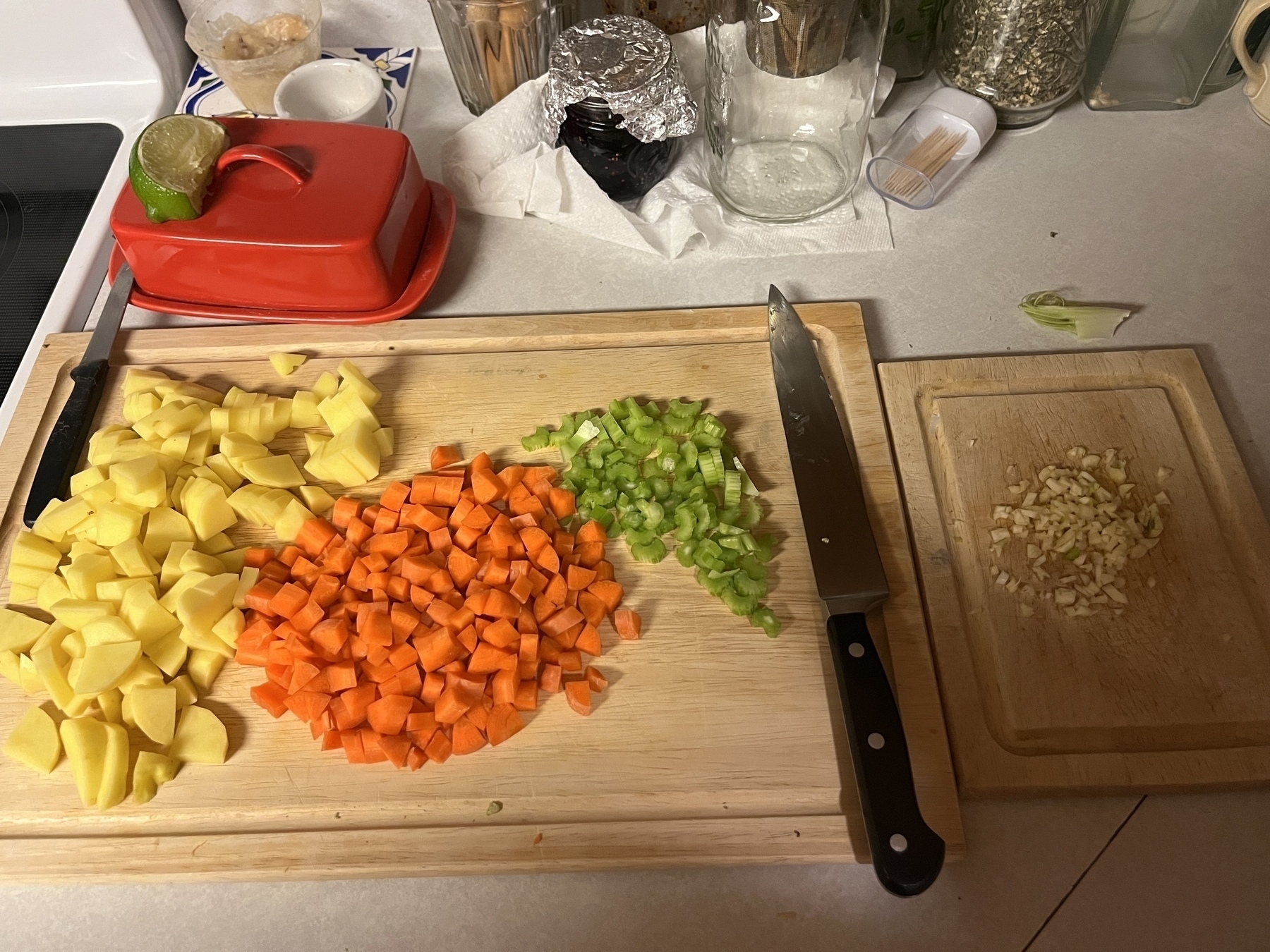 Chopped potatoes, carrots, celery, and garlic are neatly arranged on a wooden cutting board beside a knife in a kitchen setting.