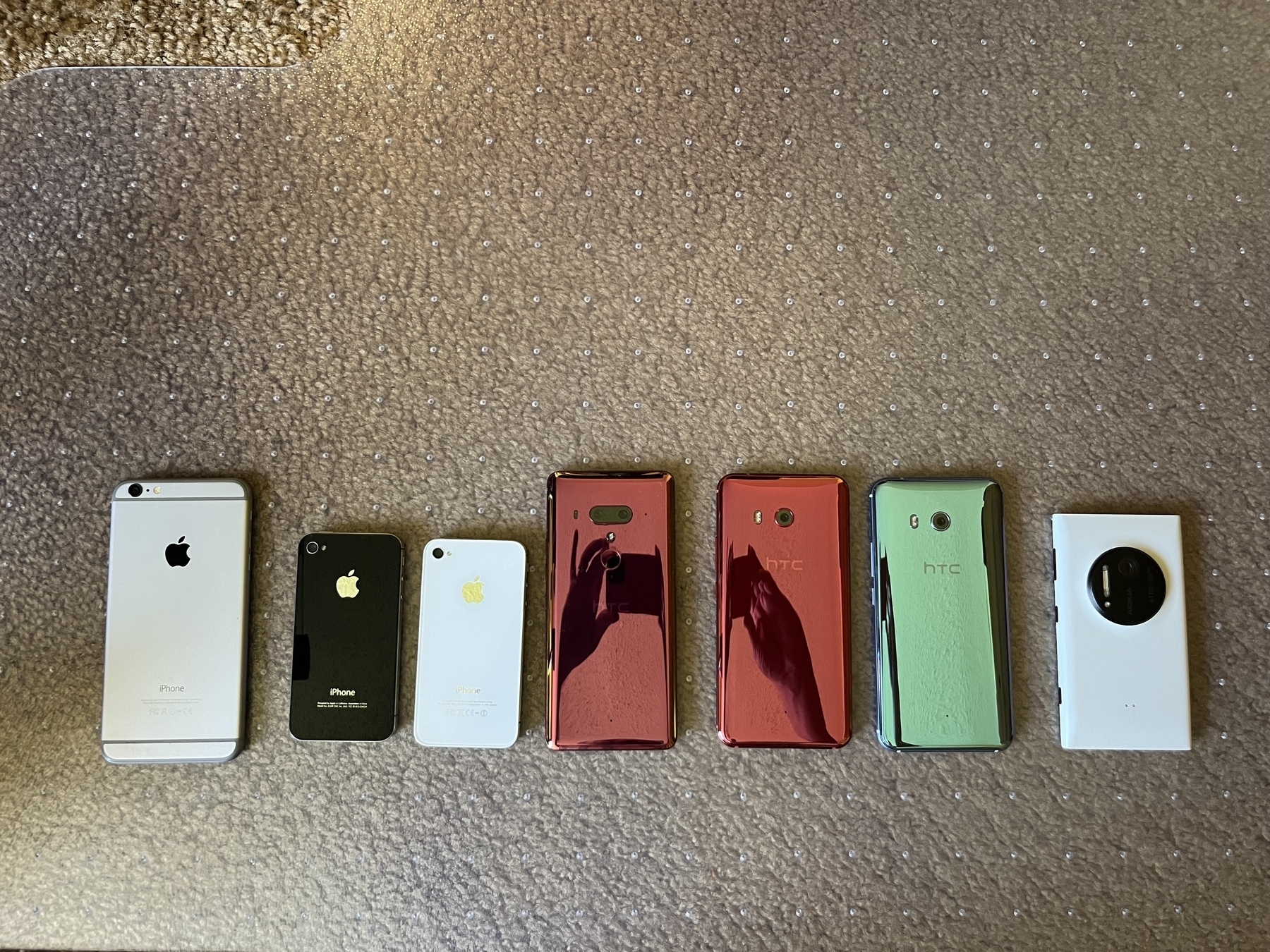 A collection of seven smartphones with different designs and colors is arranged in a line on a carpeted floor.