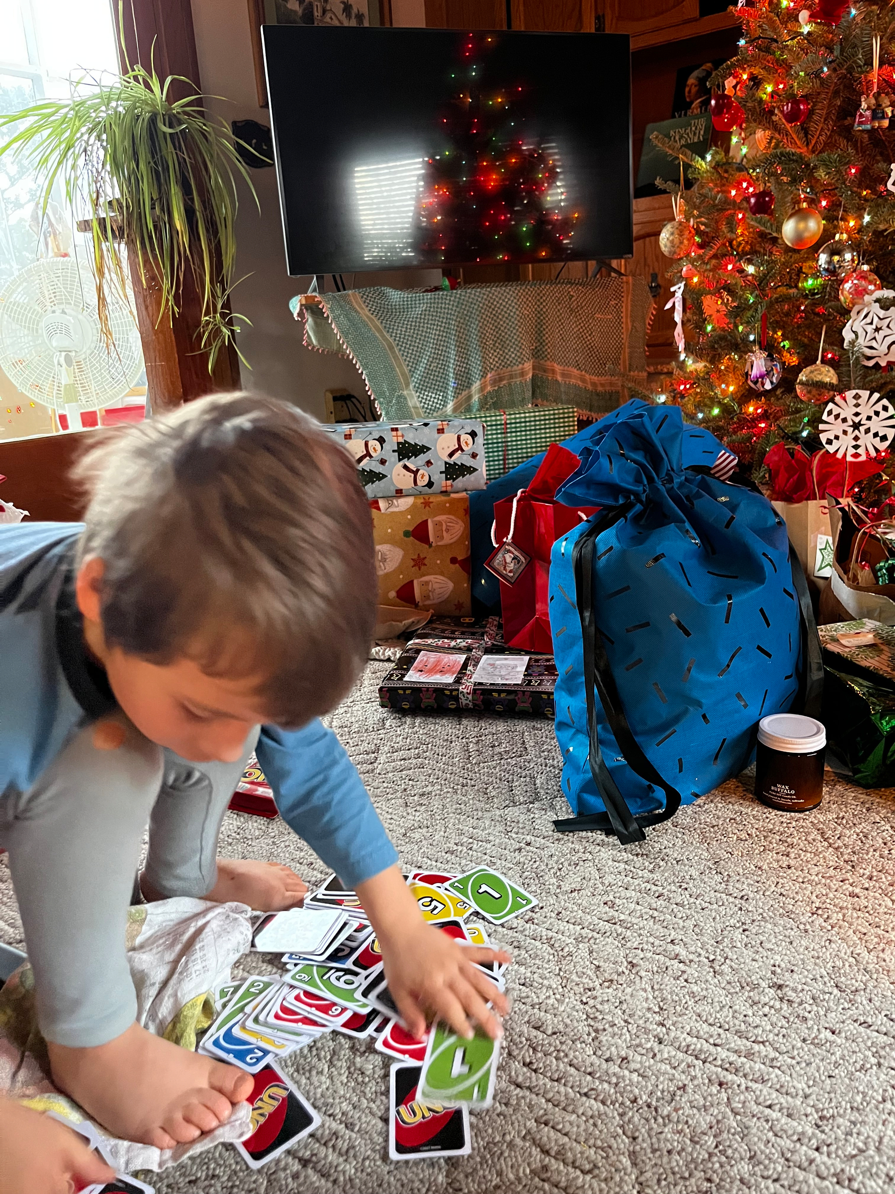 A child wearing pajamas is playing with UNO cards in front of a Christmas tree surrounded by presents.