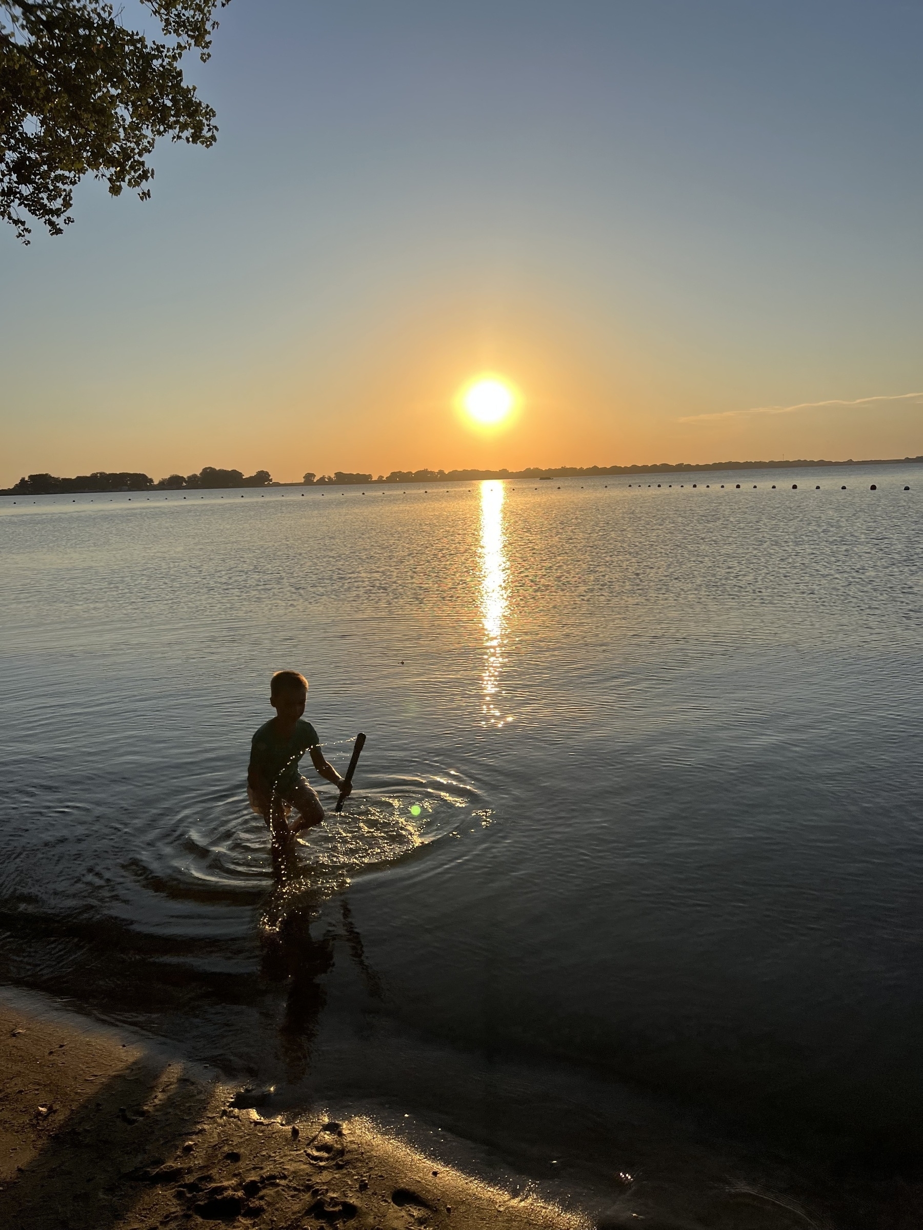 A child stands in shallow water holding a stick with a sunset reflecting on the calm lake surface.