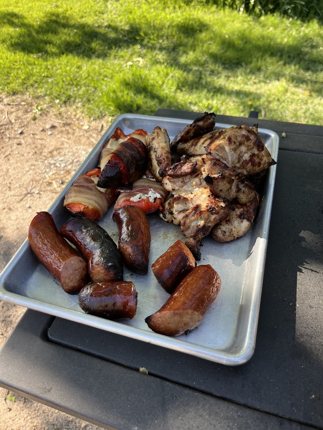 A tray is filled with grilled meats, including sausages and chicken, sitting on an outdoor table with grass in the background.
