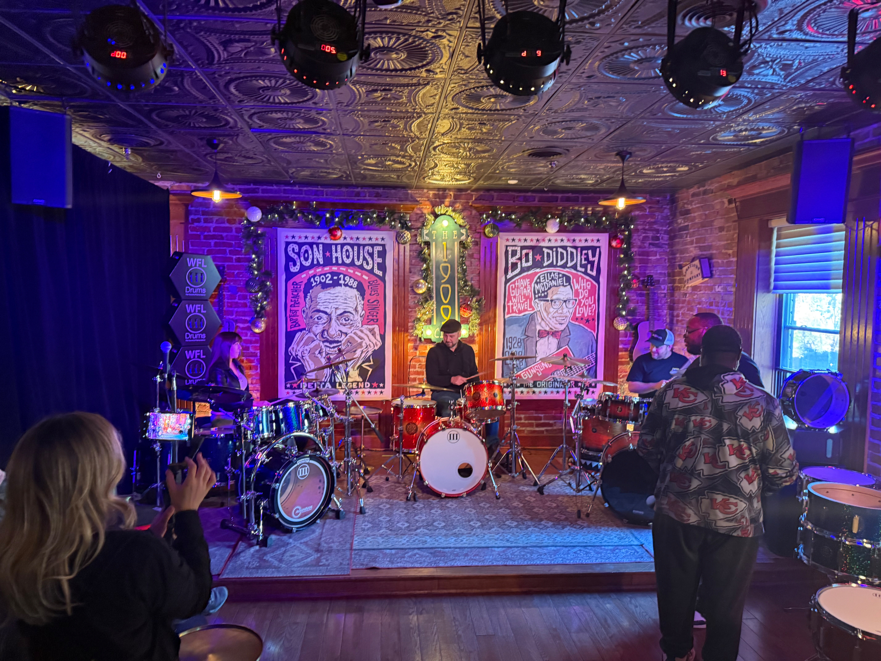 A group of musicians is playing drums on a stage adorned with vibrant posters and colorful lights, while an audience watches.