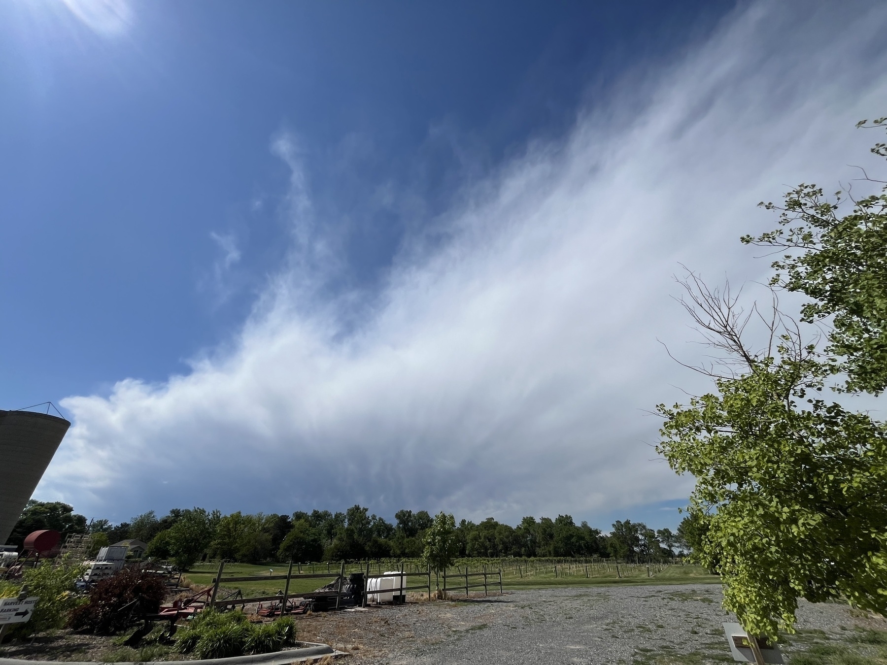 A vast sky with wispy clouds stretches over a rural landscape with scattered greenery and a gravel area.