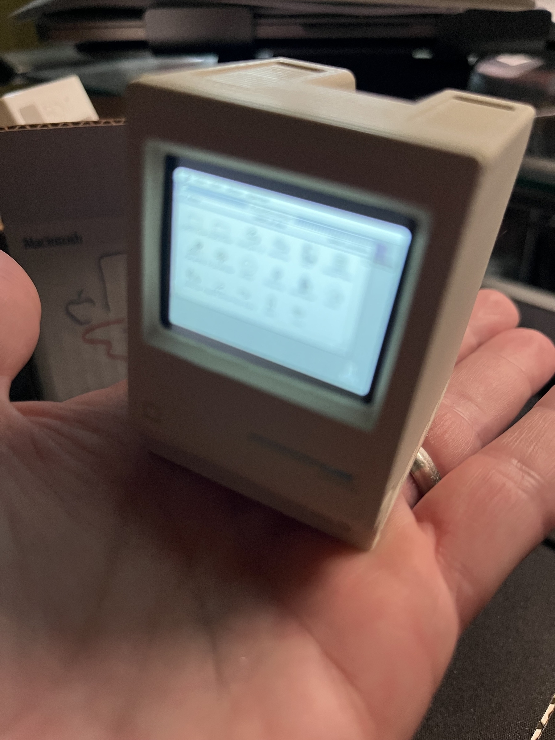 A miniature model of a classic Macintosh computer is held in a person's hand, showing a lit-up screen with icons.