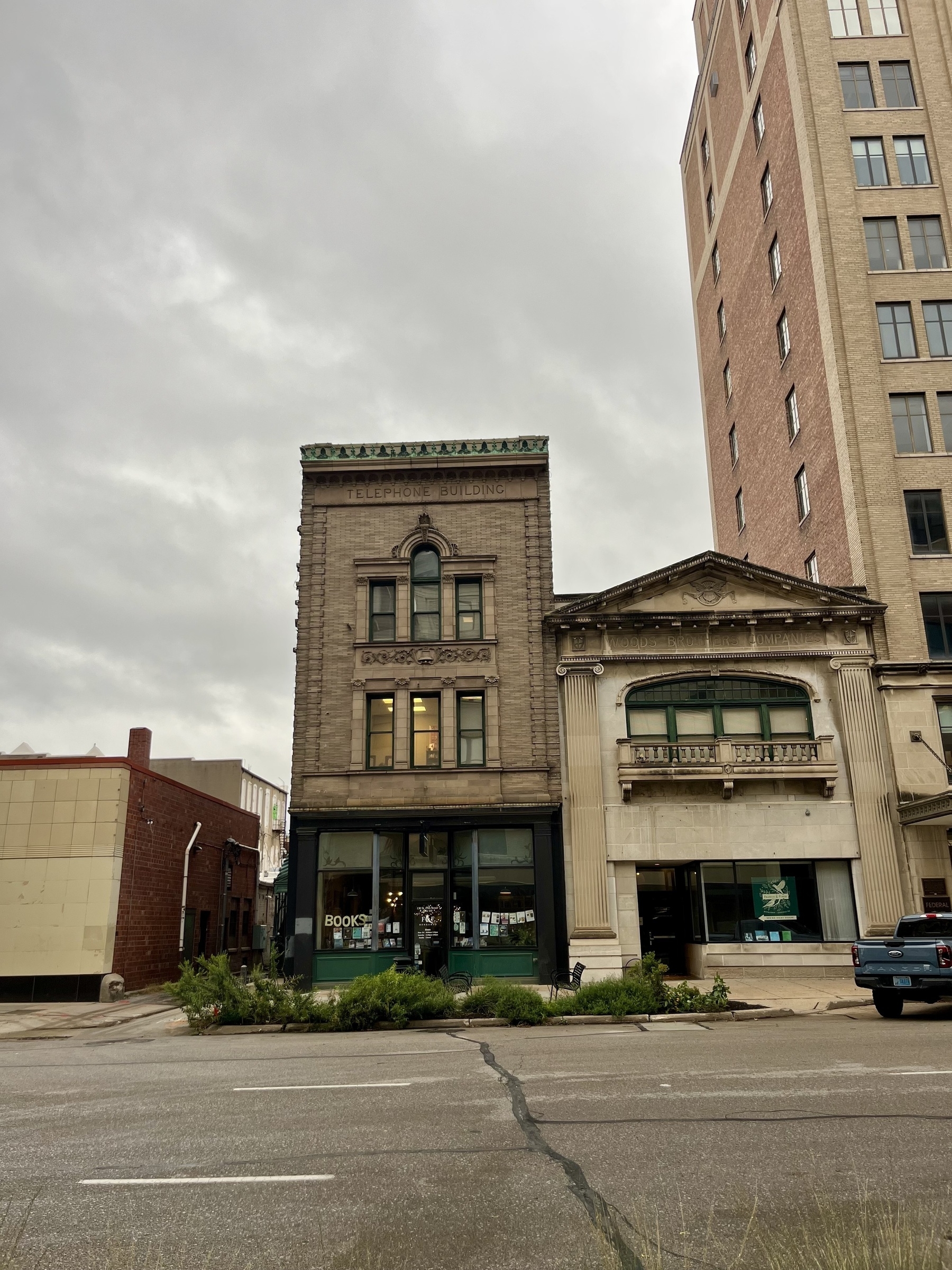 Two historical buildings with distinct architectural styles stand side by side on a cloudy day, bordered by a road and grassy patches.