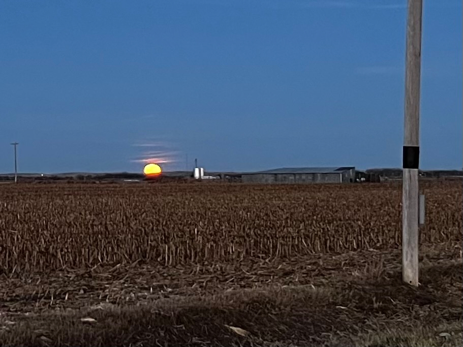 A large orange moon rises behind a flat agricultural landscape with a field, farm buildings, and power lines.