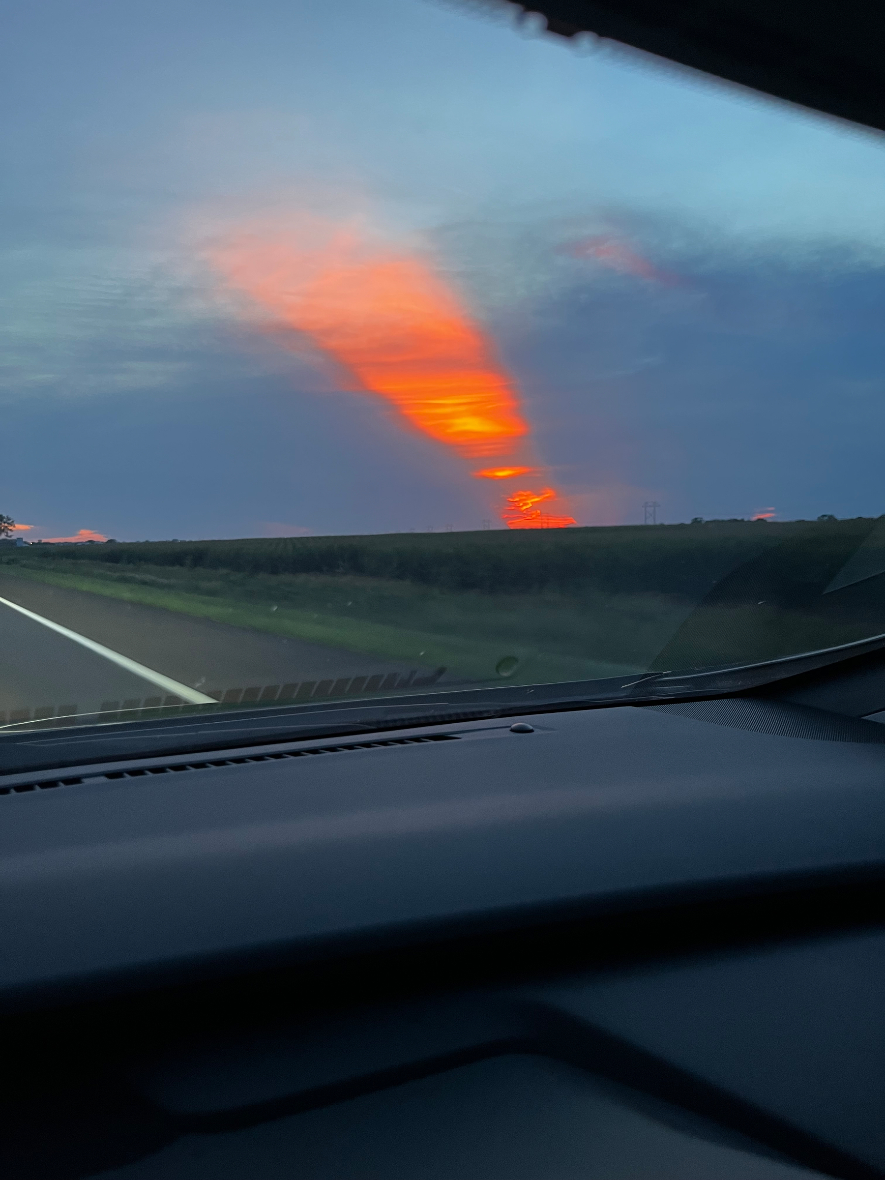 orange and pink sunset is seen through the windshield of a car driving along an open road with fields on either side.