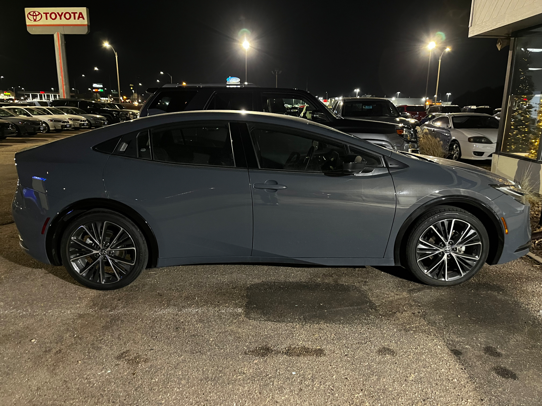 A blue modern hatchback (Toyots Prius) is parked in a car dealership lot at night, with various cars and dealership signs visible in the background.